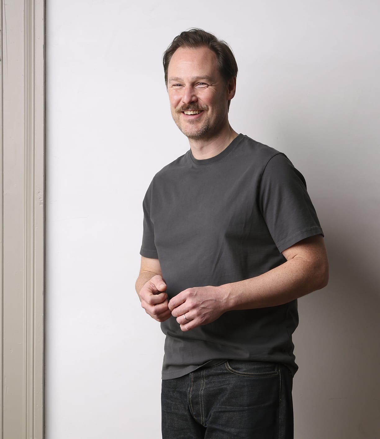 A man with light brown hair and a moustache smiles against a plain light wall, wearing Best Made Co's Standard T-Shirt in Faded Black—crafted from soft Peruvian tangüis cotton—and dark jeans, embodying a timeless staple.