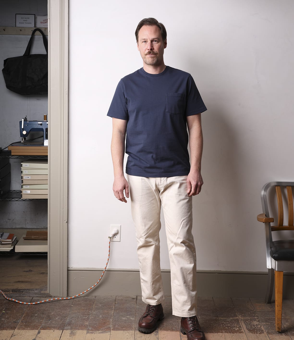 A man with short hair and a beard stands indoors on a wooden floor, wearing the Best Made Co Standard Pocket T-Shirt in navy, light-colored pants, and brown shoes. Behind him are a wooden chair, shelving, and a sewing machine.