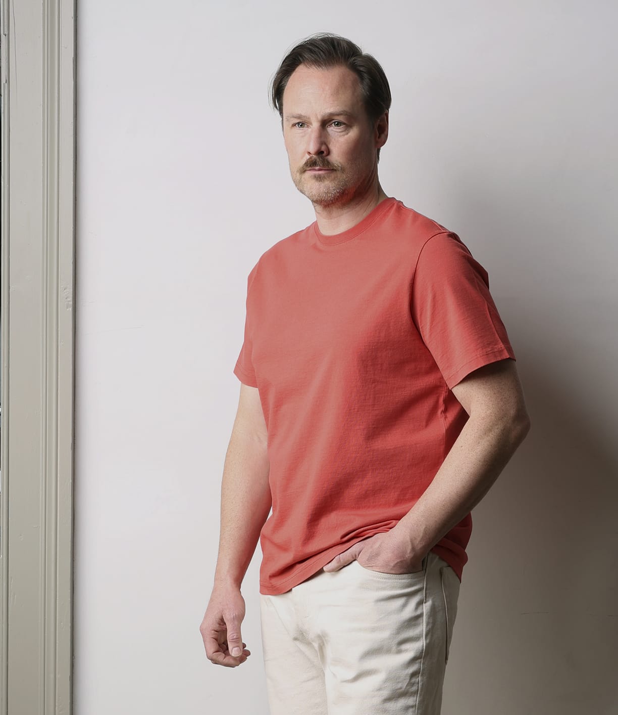 A man with light brown hair, a mustache, and stubble wears the Best Made Co Standard T-Shirt in Faded Red with light beige pants, standing indoors against a plain white wall beside a taupe door frame.