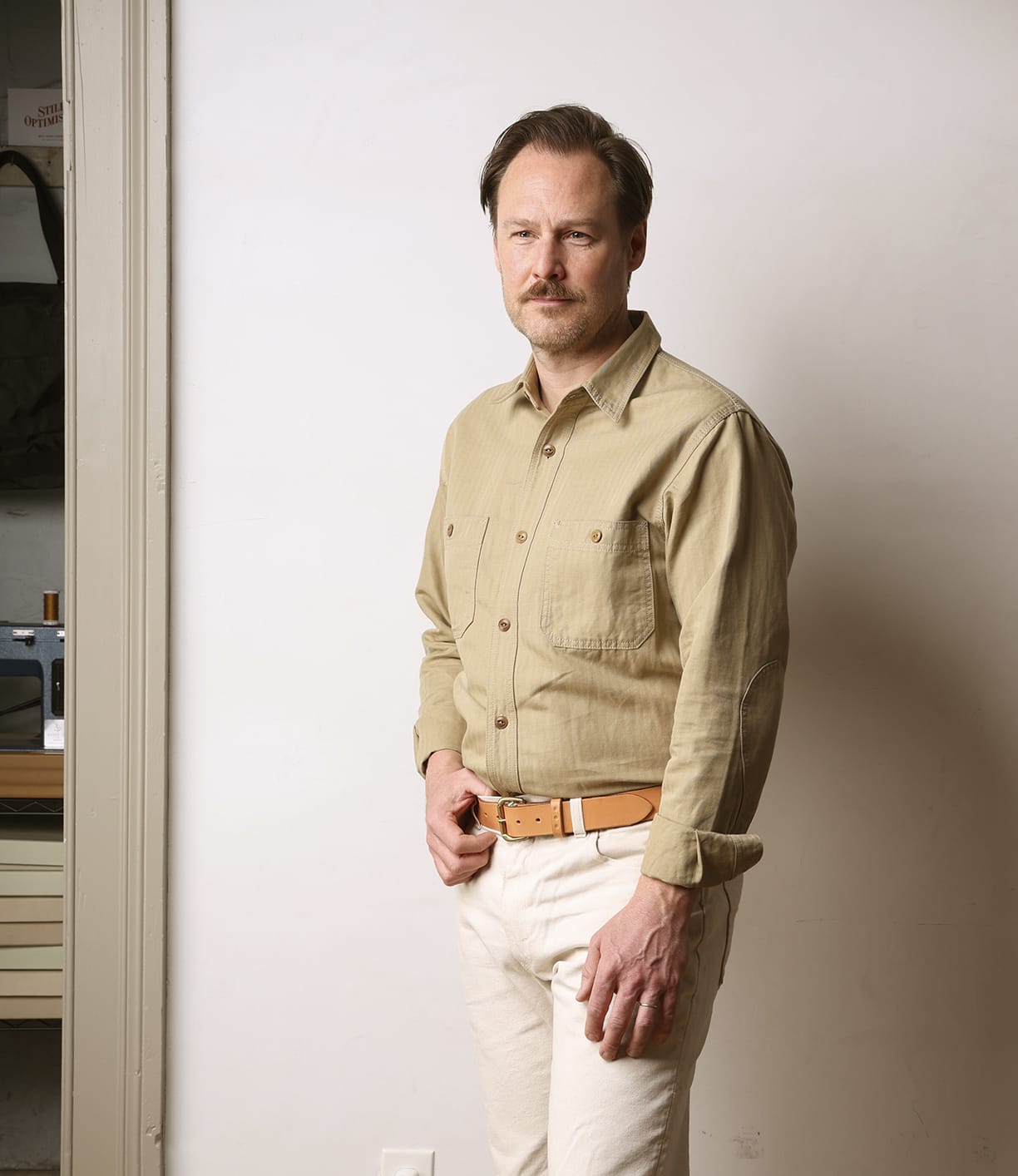 A man with light brown hair and a mustache stands indoors against a plain wall, wearing the Best Made Co. Cotton-Linen Workshirt in tan, paired with light pants and a tan belt, looking at the camera with a neutral expression.