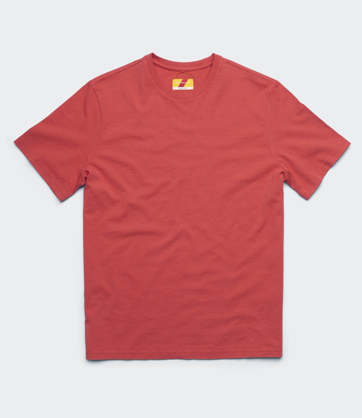 The Best Made Co Standard T-Shirt in Faded Red, a Peruvian tangüis cotton crew neck with short sleeves and garment-dyed finish, is laid flat on a light gray surface. The inner collar tag is partially visible with yellow and red details.