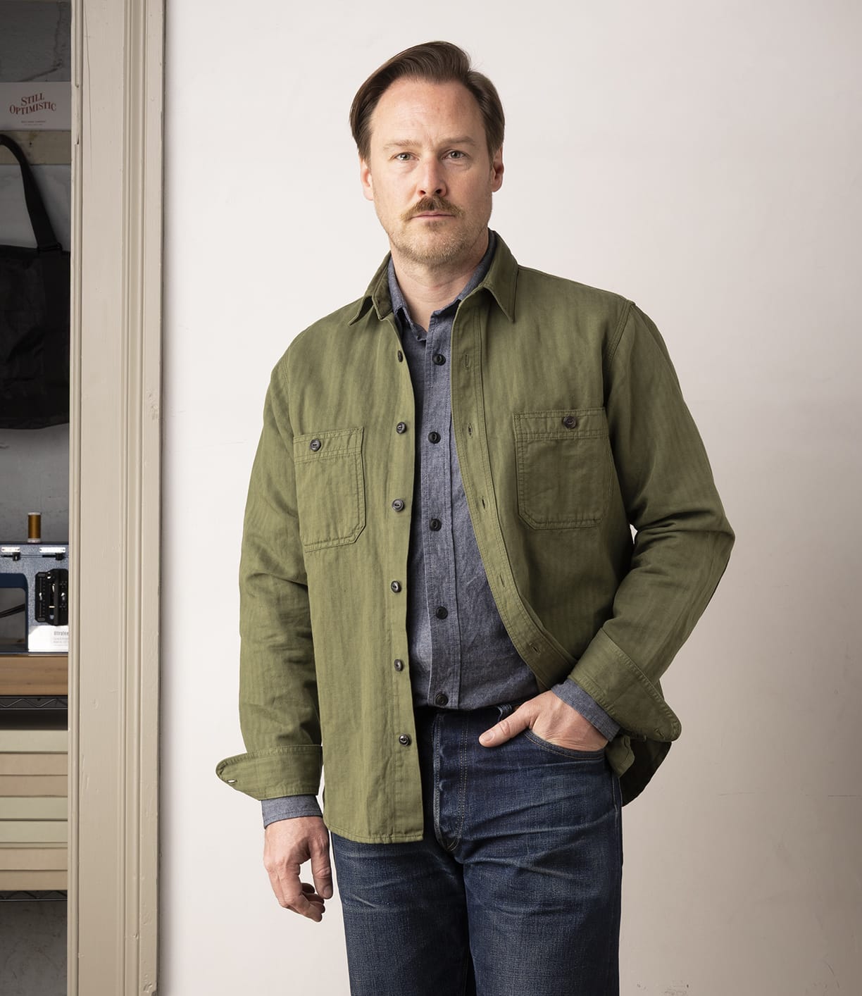 A man with light brown hair and a mustache wears the Best Made Co. Cotton-Linen Workshirt in Olive Drab over a blue-gray chambray shirt and jeans, standing indoors near a doorway with a neutral expression.