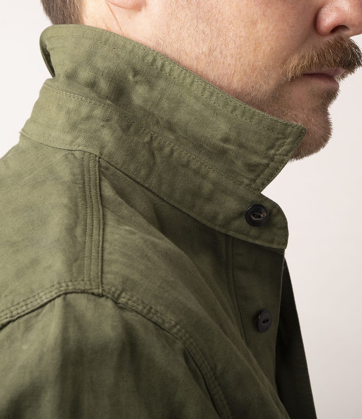 Close-up side view of a light-skinned person with a mustache wearing the Best Made Co. Cotton-Linen Workshirt in Olive Drab, collar turned up, against a plain light background.