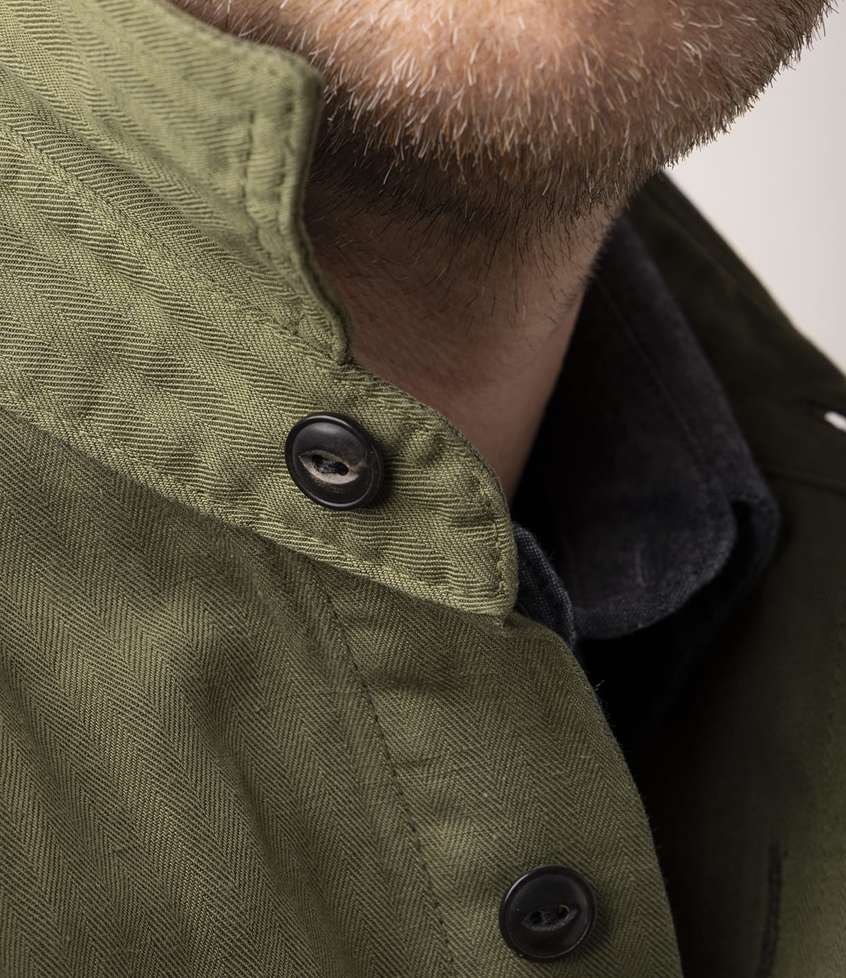 Close-up of a person’s lower face with light facial hair, wearing the Best Made Co. Cotton-Linen Workshirt in Olive Drab, featuring black buttons and a herringbone pattern, layered over a dark selvedge chambray shirt.