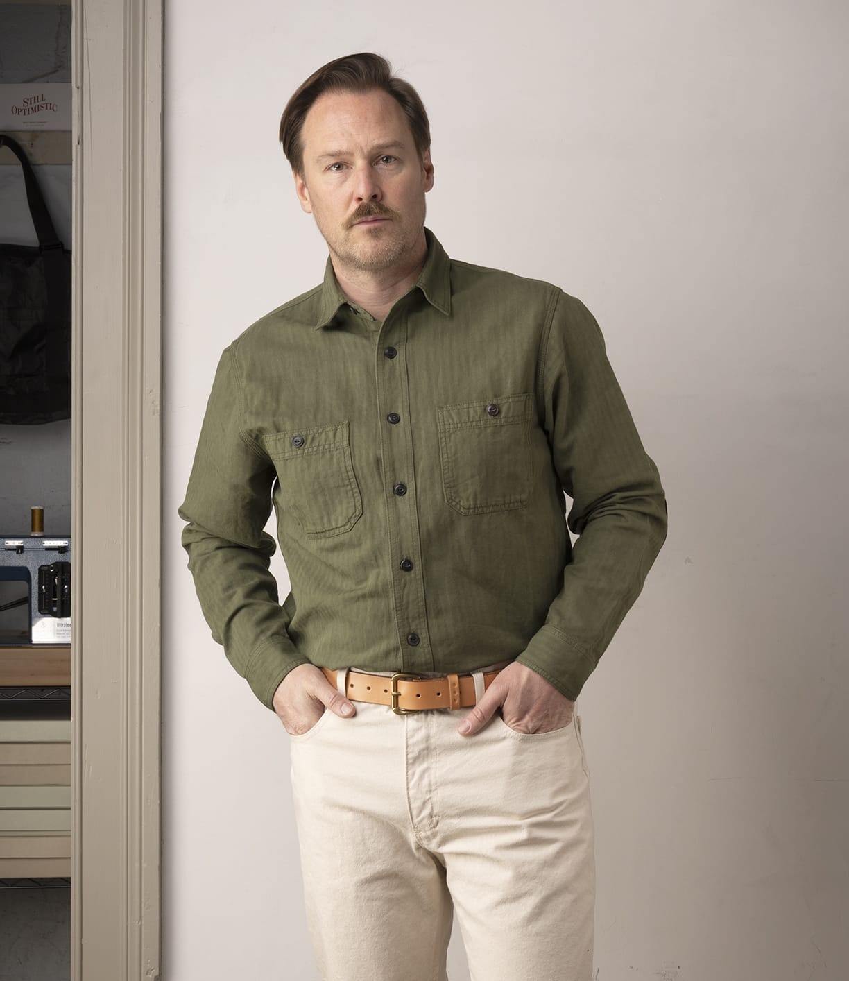 A man with light brown hair and a mustache stands indoors wearing the Best Made Co. Cotton-Linen Workshirt in Olive Drab, beige pants, and a tan belt. He has his hands in his pockets and looks at the camera with a neutral expression.