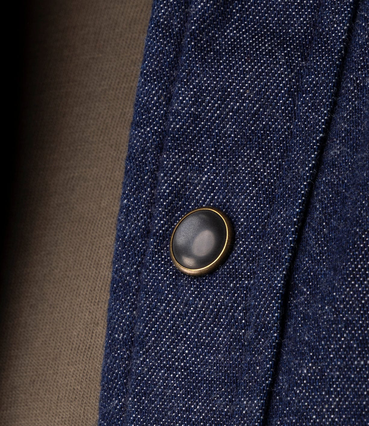 Close-up of the Fukuyama Snap-Front Shirt by Best Made Co, featuring a dark metal snap button with a gold rim on indigo-dyed denim, with brown fabric in the background.