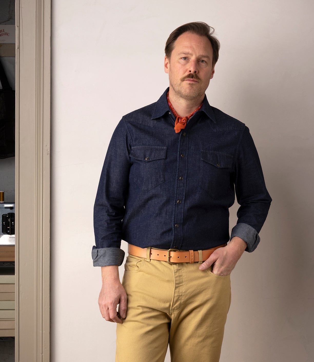 A man with light brown hair and a mustache stands indoors against a plain wall, wearing the Best Made Co Fukuyama Snap-Front Shirt, tan pants, a tan belt, and a red neckerchief. One hand is in his pocket as he looks at the camera.