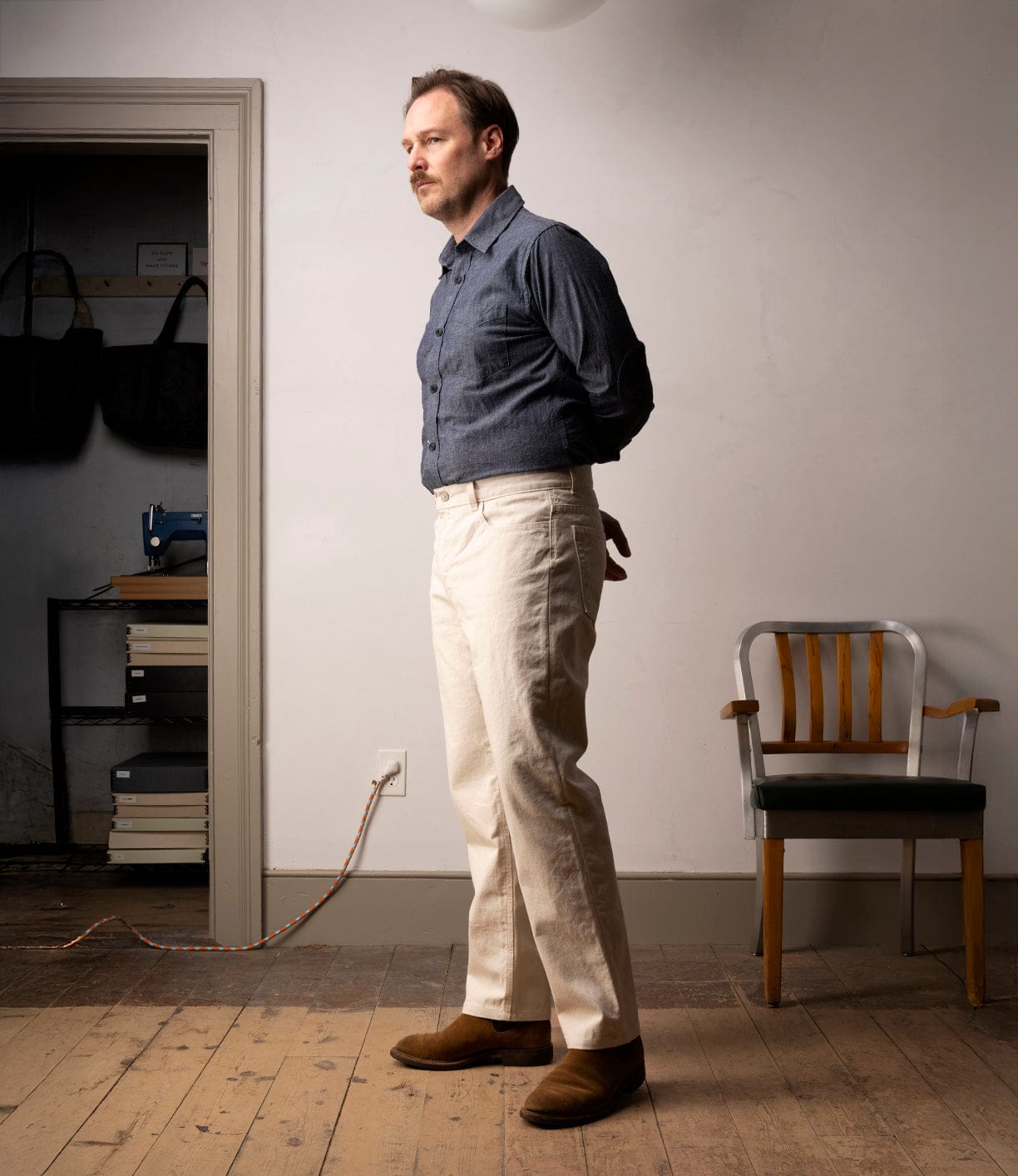 A light-skinned man in a blue shirt and Best Made Co Standard 5-Pocket Pant M2 Undyed stands indoors with hands behind his back. Behind him are a wooden chair and a shelf holding bags and books.