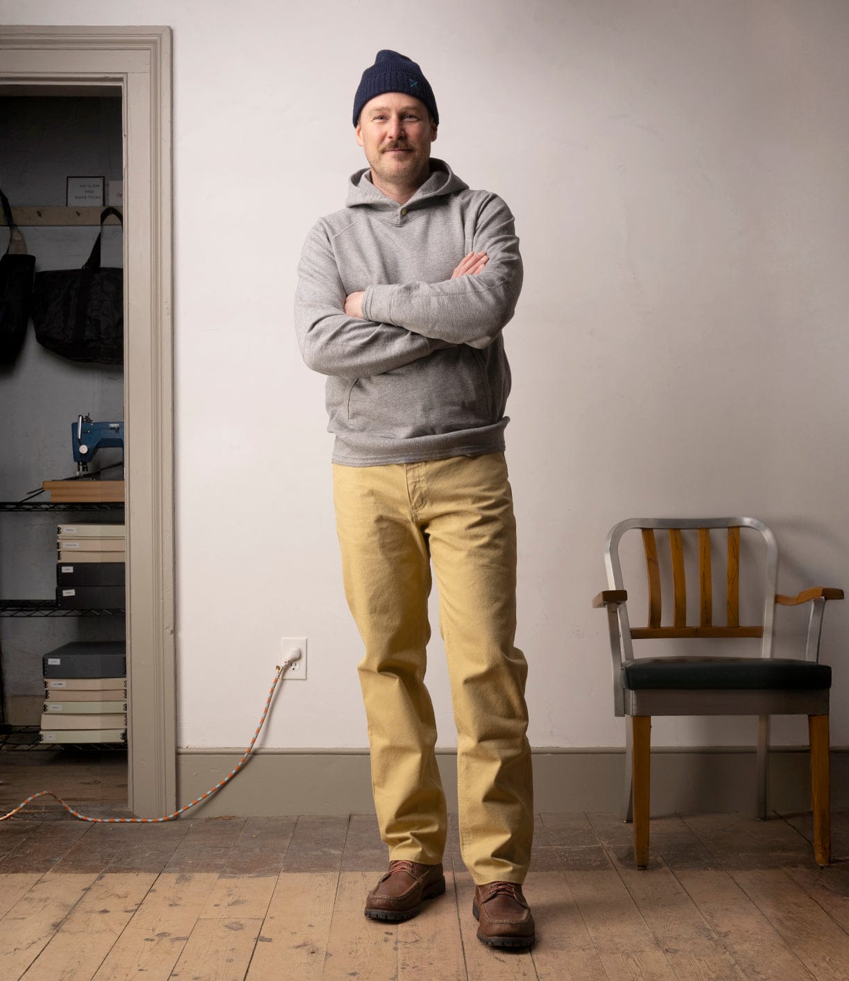 A man with a mustache, navy beanie, gray hoodie, Best Made Co Standard 5-Pocket Pant M2 Khaki, and brown shoes stands arms crossed in a room with wooden floors beside a gray chair and a shelf with bags and books.