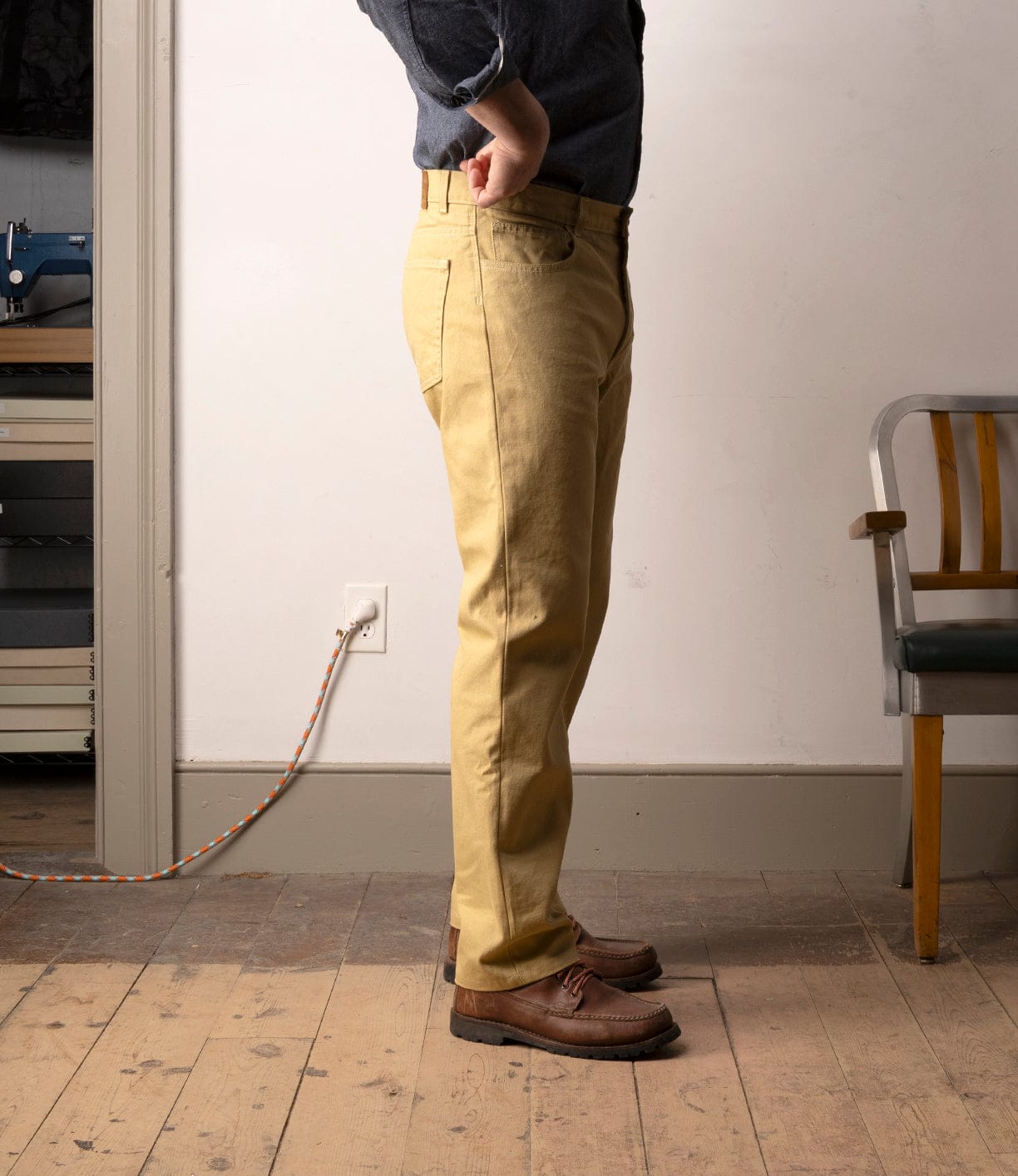 A person in a blue shirt, Best Made Co Standard 5-Pocket Pant M2 Khaki, and brown shoes stands sideways on a wooden floor near a gray and wood chair, with an electrical cord plugged into the wall visible.