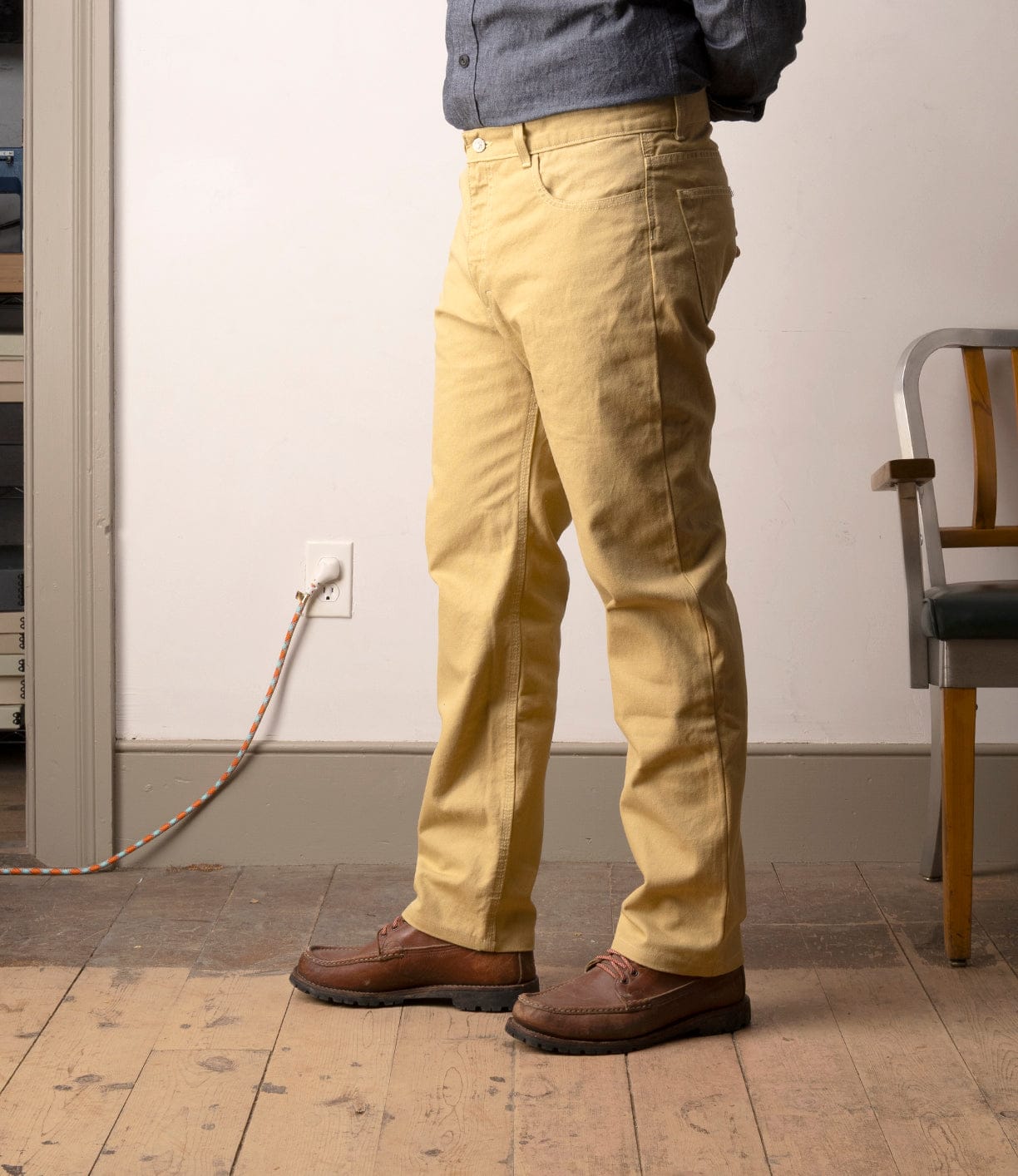 A person’s lower body is shown indoors, wearing Best Made Co’s Standard 5-Pocket Pant M2 Khaki, a gray shirt, and brown shoes; they stand on a wooden floor near a power outlet and a chair.