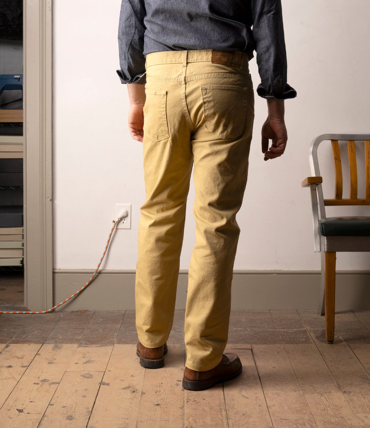 A person stands indoors facing away, wearing Best Made Co’s Standard 5-Pocket Pant M2 Khaki, brown shoes, and a blue shirt. To the right is a wooden chair; to the left, a power cord is plugged in. The floor is worn wood.