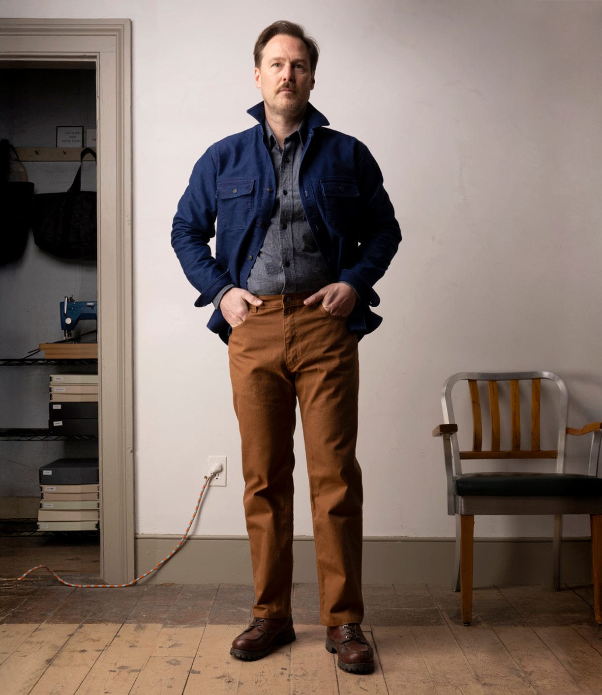 A man stands indoors on a wooden floor, wearing a blue jacket, gray shirt, and Best Made Co Standard 5-Pocket Pant M2 Field Brown with brown shoes. He is between a wooden chair and shelves with books and a bag; a cord runs along the wall.