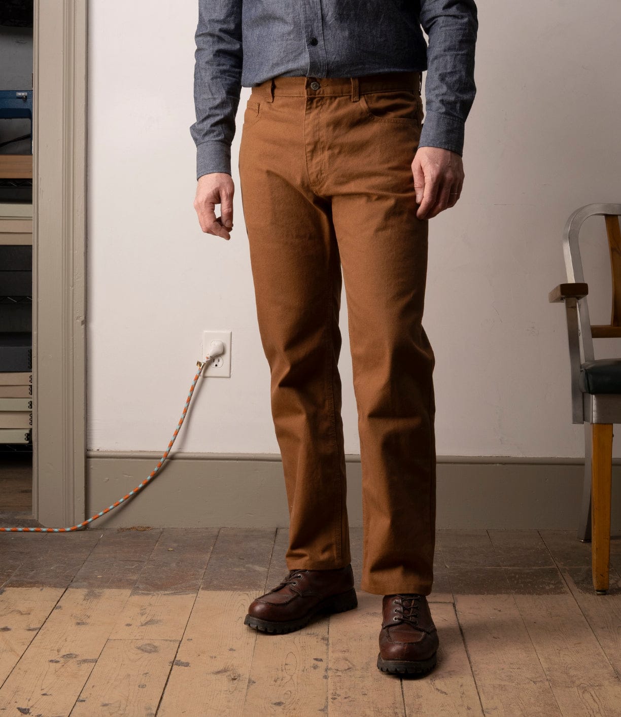 A person in a blue button-up shirt, brown shoes, and Best Made Co’s Standard 5-Pocket Pant M2 Field Brown stands indoors on a wooden floor near a power outlet and an orange-and-white extension cord.