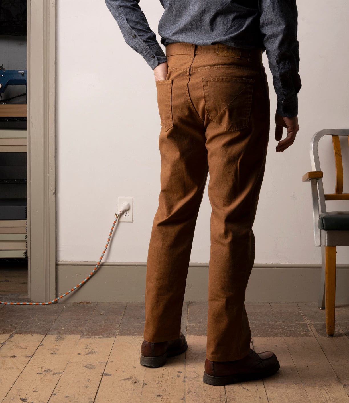 A person wears Best Made Co Standard 5-Pocket Pant M2 Field Brown, a blue long-sleeve shirt, and brown shoes while standing indoors on a wooden floor. A striped cord is plugged into the wall and a chair is partly visible on the right.