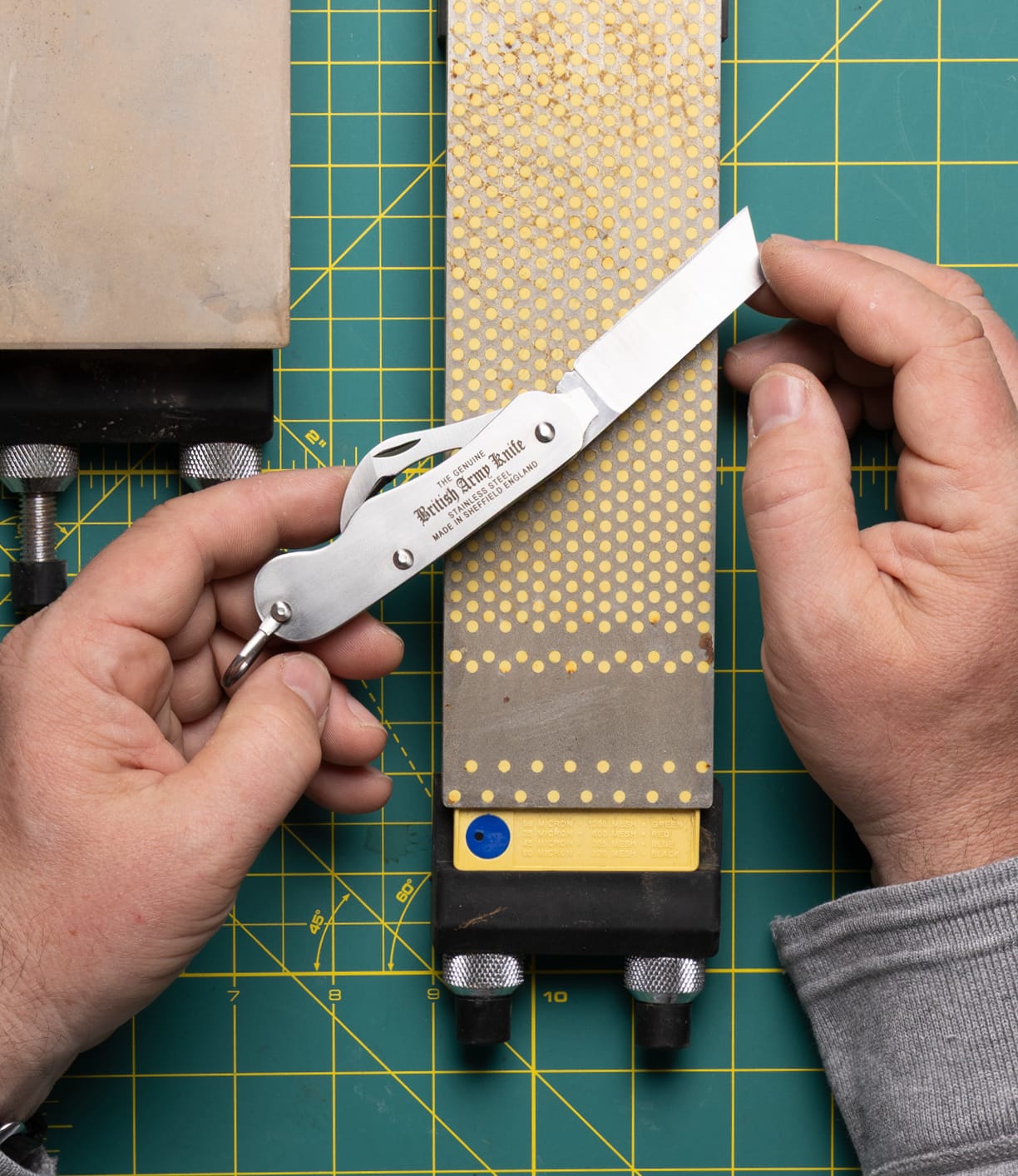 A person sharpens the Best Made Co. British Army Knife on a rectangular stone, holding the utility knife at an angle with both hands over a gridded cutting mat.