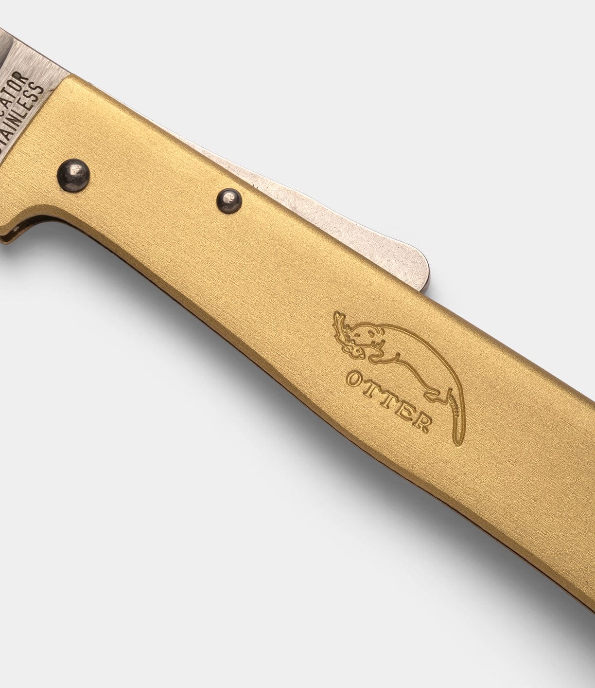 Close-up of a brass-handled Brass Mercator folding knife by Best Made Co. The handle features a metal rivet and engraved otter above "OTTER." The partially visible carbon steel blade is stamped with text.