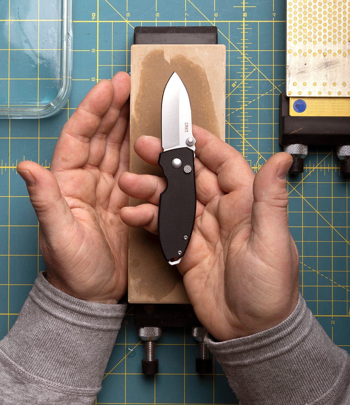 A person holds a Best Made Co CRKT Squid knife with a D2 steel blade and black handle above a sharpening stone, both hands around the knife, on a cutting mat.