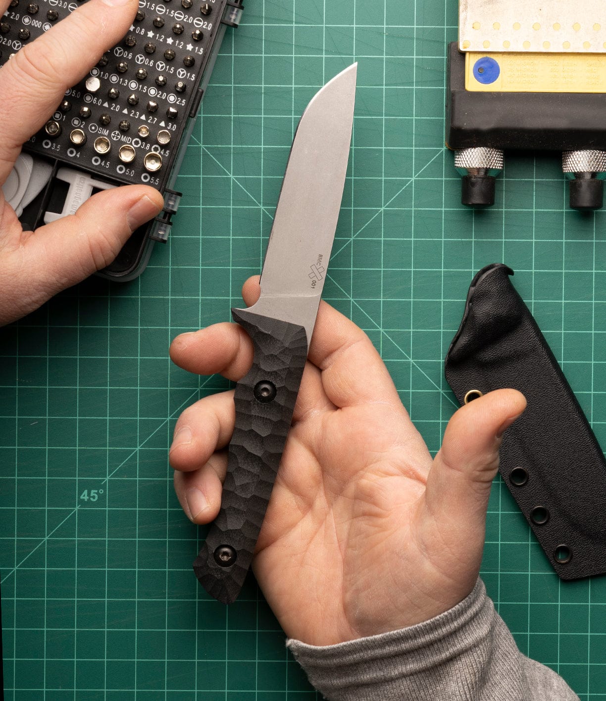 Someone holds the Best Made Co NWBW Field Knife (BMC Edition) with a textured black handle made from American D2 tool steel above a green cutting mat, alongside a tool kit, knife sheath, and sharpening block.