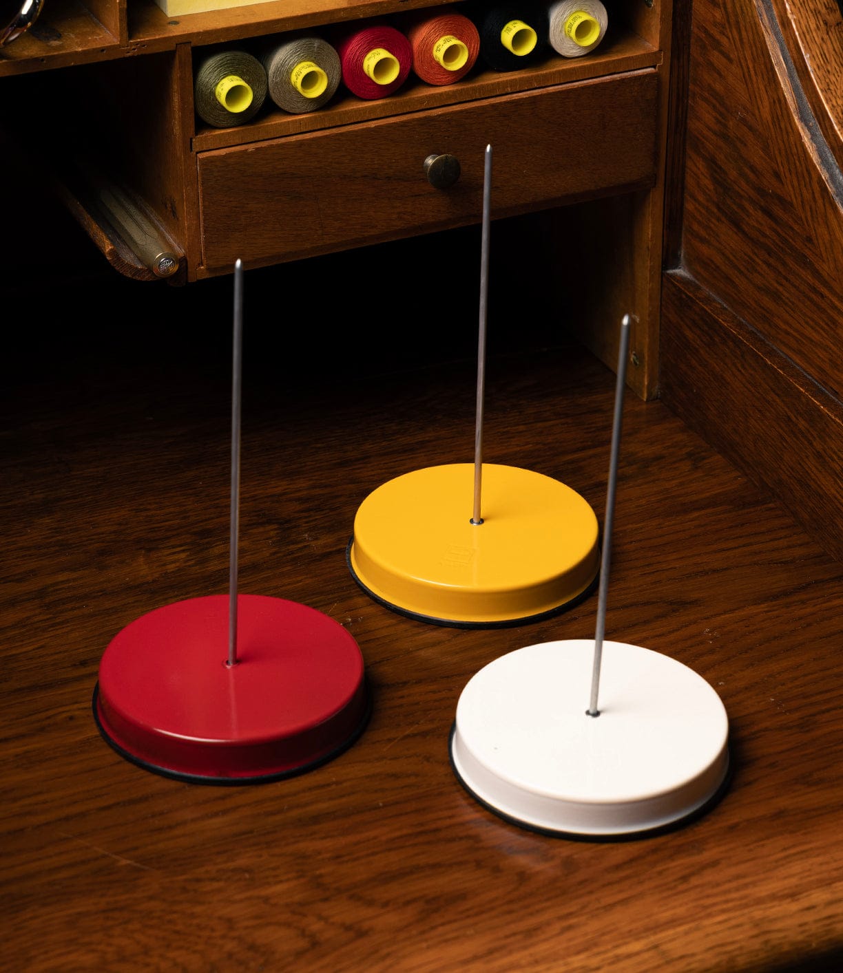 Three round powder-coated steel Italian Receipt Spikes by Best Made Co. in red, yellow, and white sit on a wooden desk. Open drawers behind them reveal matching spools of thread, creating a vibrant organizer setup.