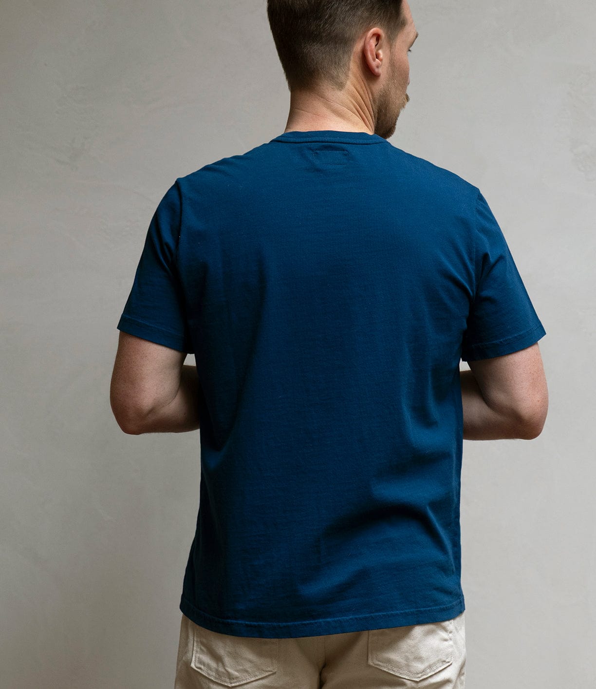 A man stands with his back to the camera, wearing Best Made Co’s Pocket T-Shirt in Haze Gray—crafted from soft Peruvian tangüis cotton—and light-colored shorts against a plain, light background.