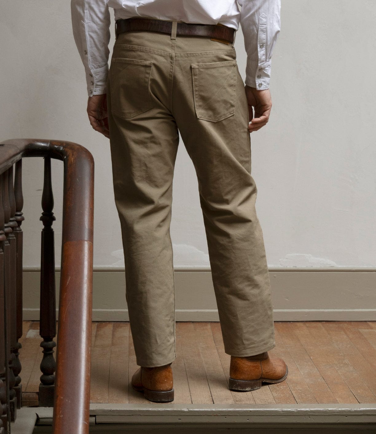 A person stands indoors on a wooden floor next to a staircase railing, facing away from the camera, wearing a white shirt, brown belt, brown shoes, and Best Made Co Canvas 5-Pocket Pant Mark 2 - Undyed.