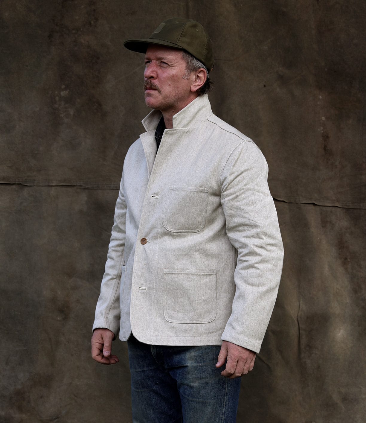 A man with a mustache in a Best Made Co Selvedge Beige Studio Jacket, blue jeans, and a green cap stands before a textured brown background, glancing aside—a nod to timeless American craftsmanship.