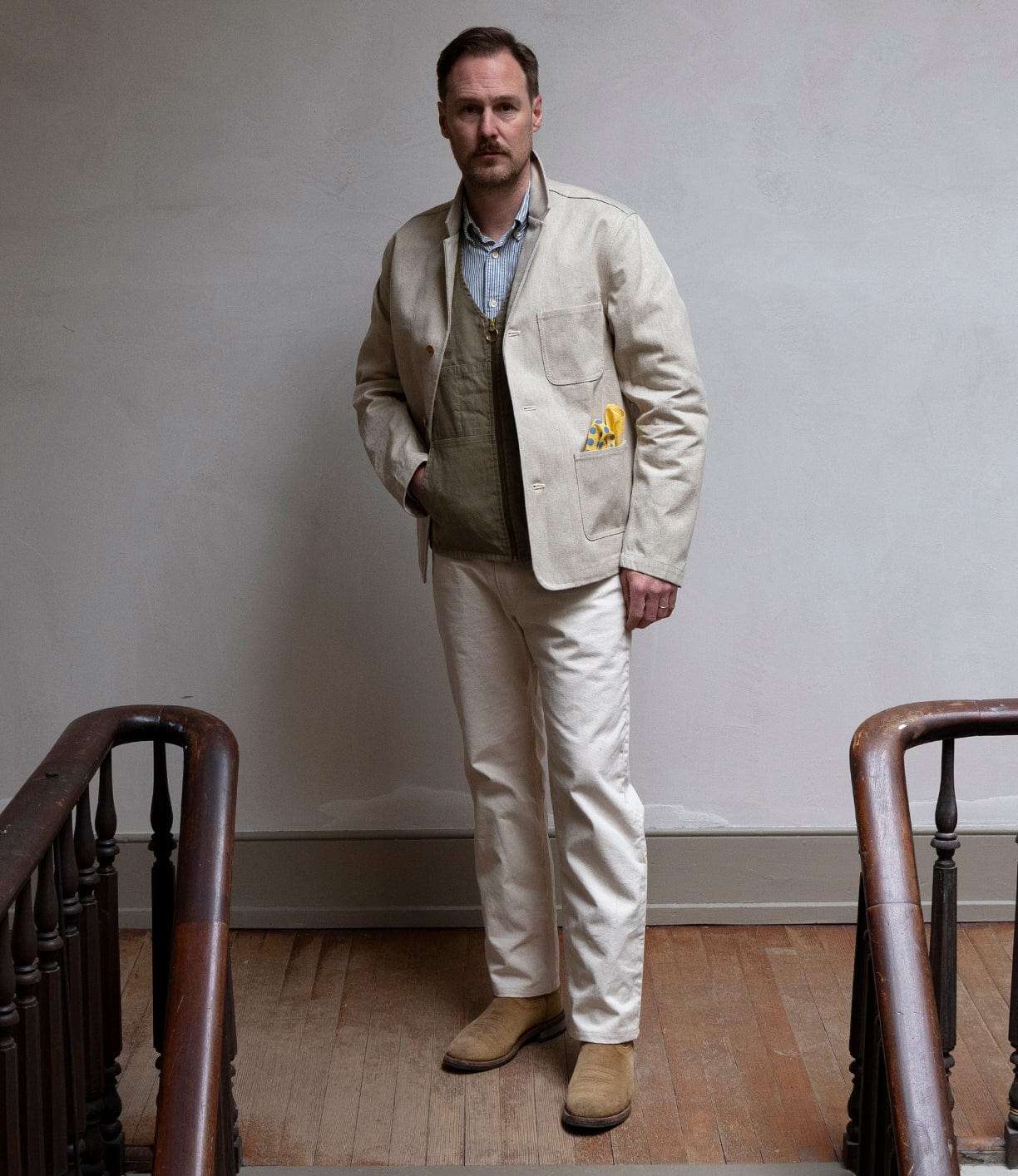 A man with a mustache stands indoors between two wooden railings, posing with one hand in his pocket while wearing the Best Made Co Selvedge Beige Studio Jacket, cream pants, an olive green vest, light blue shirt, and tan suede boots.