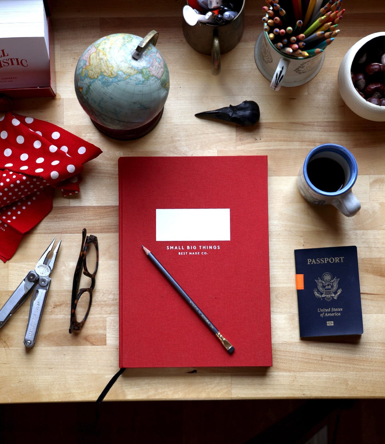 A wooden desk with a Best Made Co. S.B.T. Max Cloth-Bound Notebook, pencil, multitool, glasses, globe, coffee mug, U.S. passport, colored pencils, red polka dot cloth, black bird figurine, and a bowl of chestnuts.