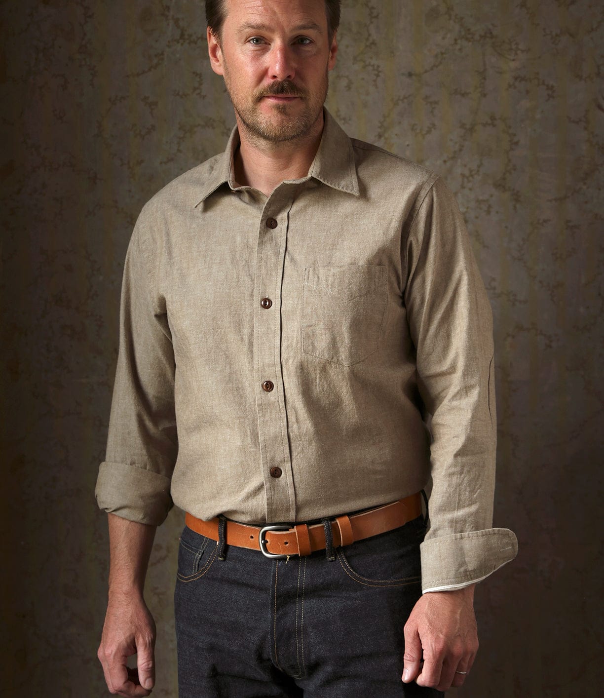 A man stands against a patterned background, wearing the Best Made Co. Cotton/Linen Herringbone Workshirt in tan with rolled-up sleeves, a tan leather belt, and dark jeans. He has short hair and a beard, looking slightly toward the camera.