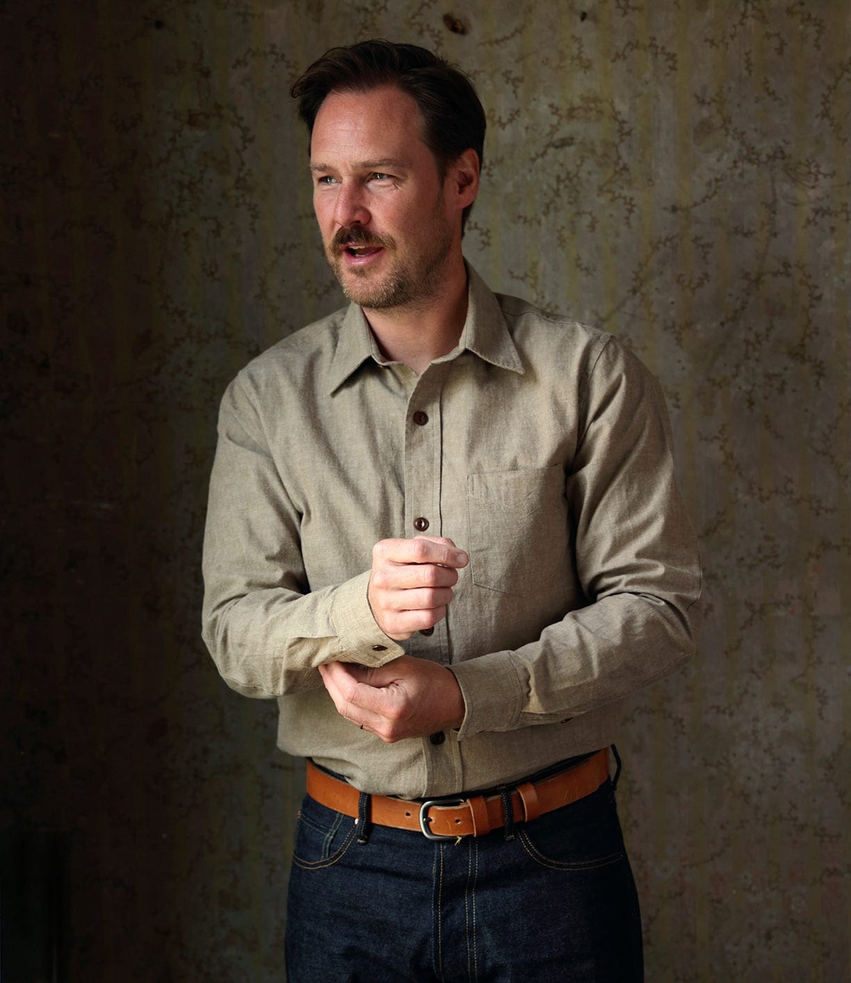 A man with short brown hair and a mustache stands against a textured wall, wearing the Best Made Co. Cotton/Linen Herringbone Workshirt in tan tucked into dark jeans with a tan belt, adjusting his left cuff with his right hand.