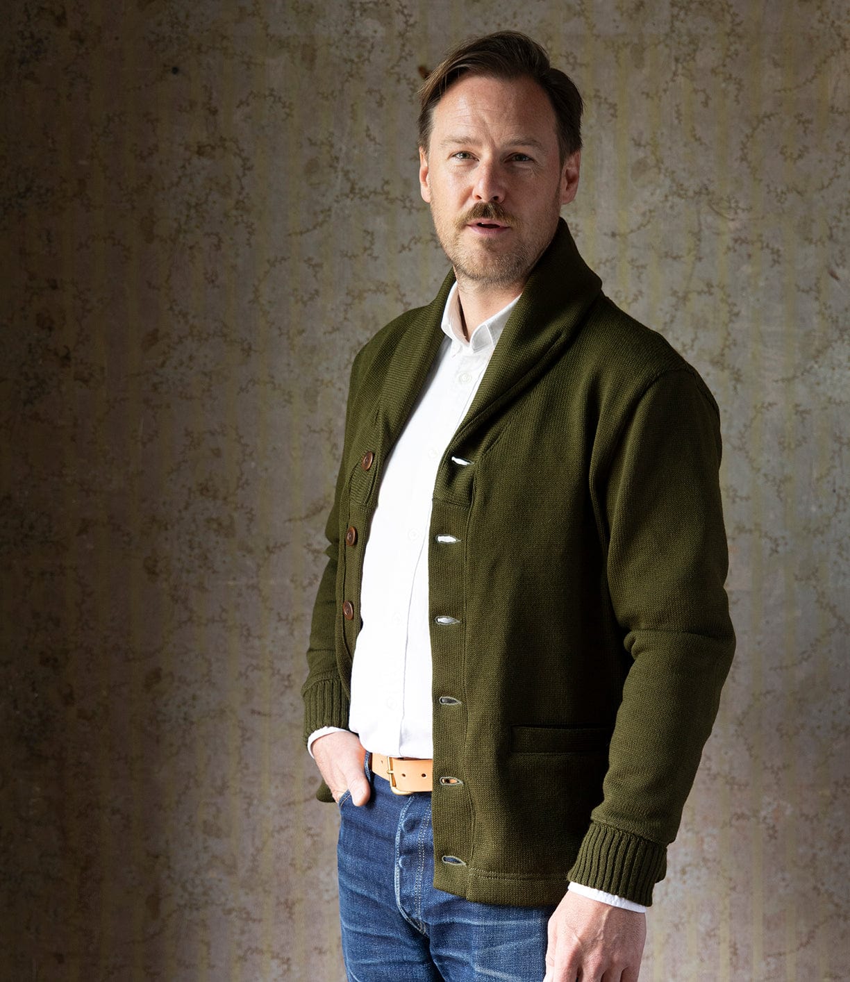 A man with light brown hair, a mustache, and a trimmed beard stands against a neutral wall, wearing the Best Made Co. Dehen Shawl Neck Sweater in Moss Green over a white shirt, blue jeans, and a tan belt, looking slightly toward the camera.