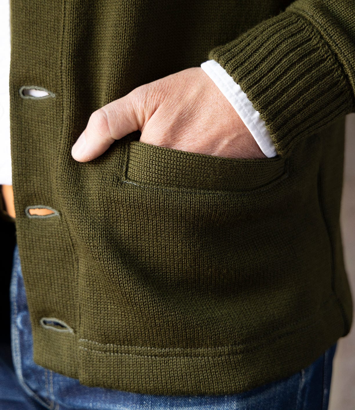 A person wears the Best Made Co. Dehen Shawl Neck Sweater in Moss Green, unbuttoned to show a white shirt and blue jeans, with their right hand in a pocket—a nod to classic American workwear style.