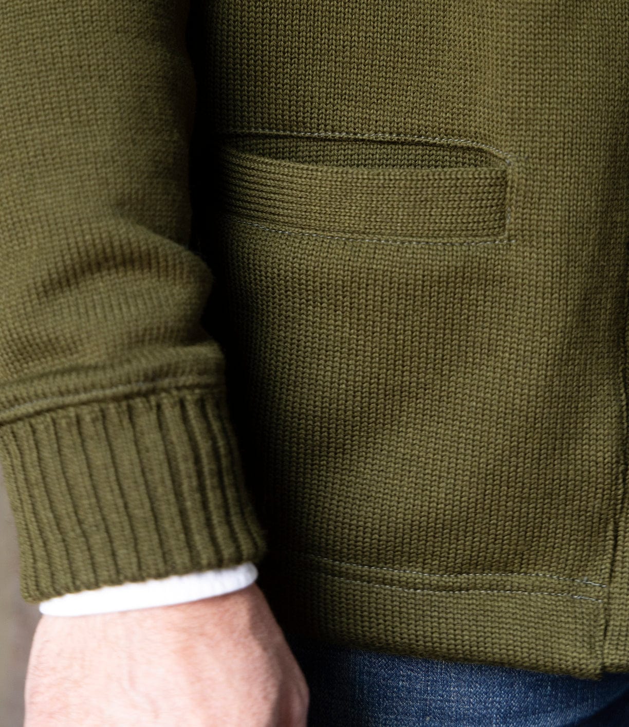 A close-up of a person’s arm and hand wearing the Best Made Co. Dehen Shawl Neck Sweater in Moss Green, with a visible pocket, cuff, white shirt sleeve, and blue jeans. Inspired by classic American workwear.