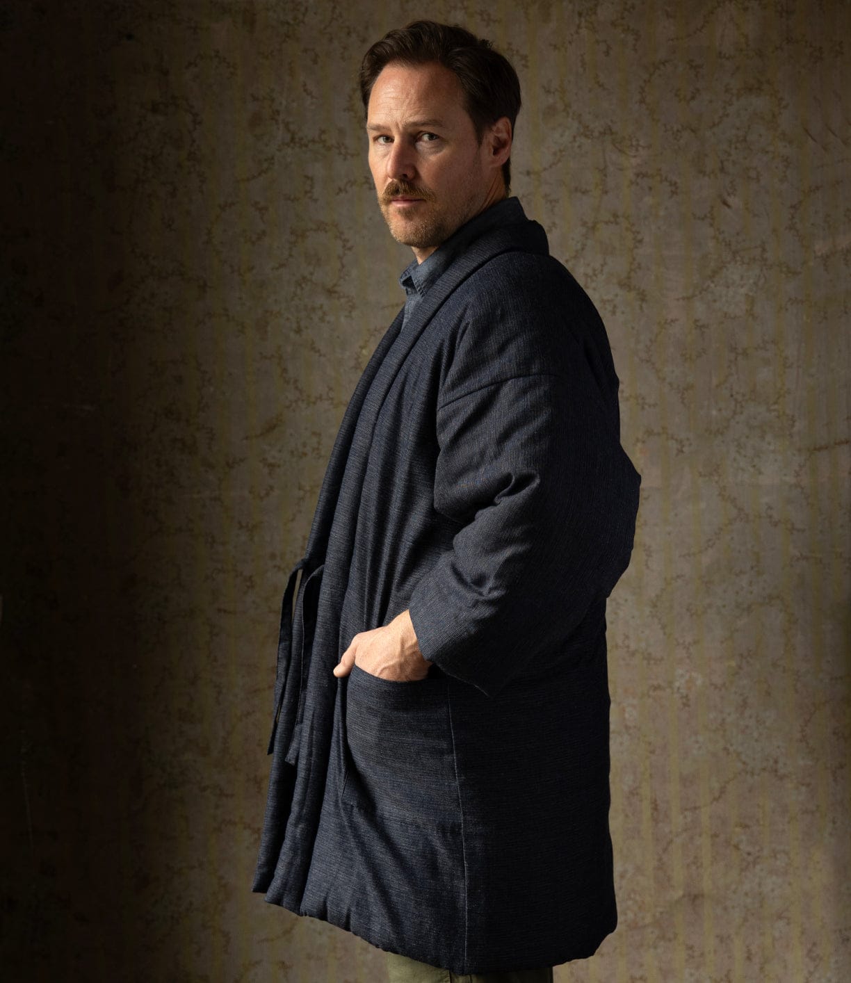 A man with short brown hair and a mustache stands sideways, looking at the camera. He wears the Best Made Co. Quilted Hanten Jacket over a shirt, inspired by Japanese workwear, with one hand in his pocket against a softly lit textured background.