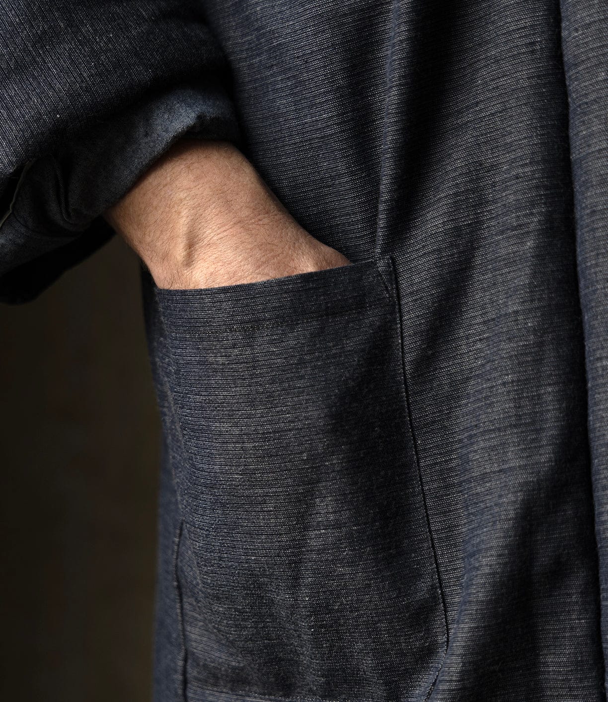 A hand rests in the pocket of a dark, textured Quilted Hanten Jacket by Best Made Co., its sleeve casually rolled up to highlight the timeless appeal of classic Japanese workwear.