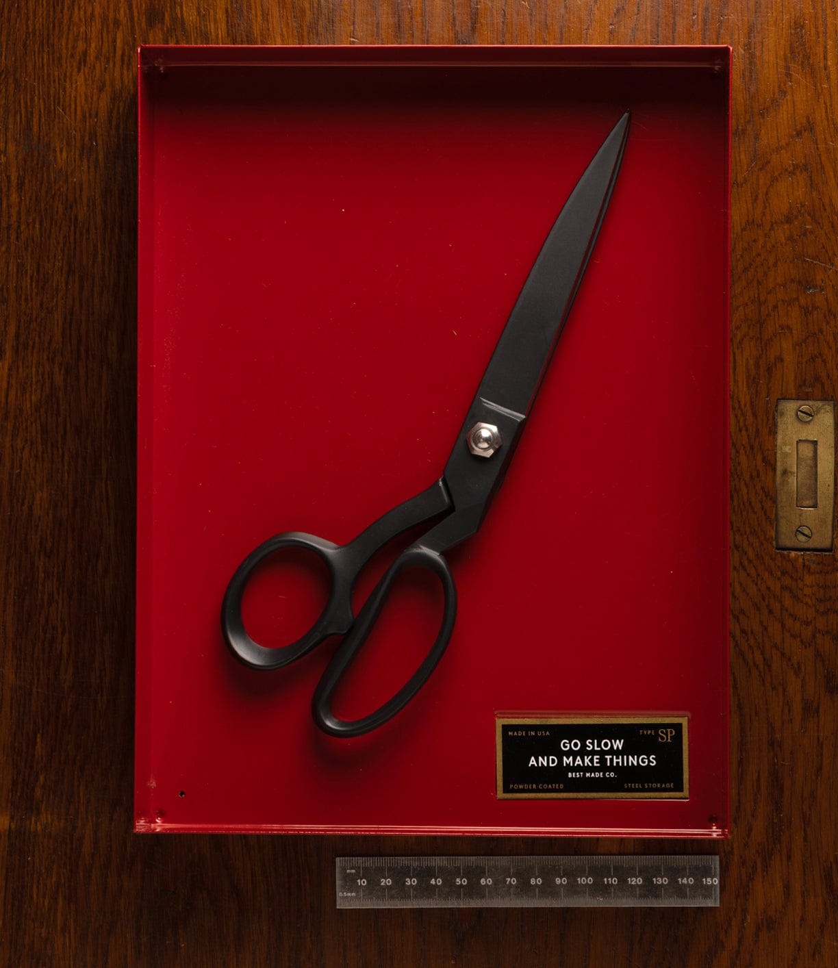 A pair of Best Made Co Kevlar Shears rests inside a red shadow box on a wooden surface, beside a transparent ruler and a plaque that reads "Go slow and make things.