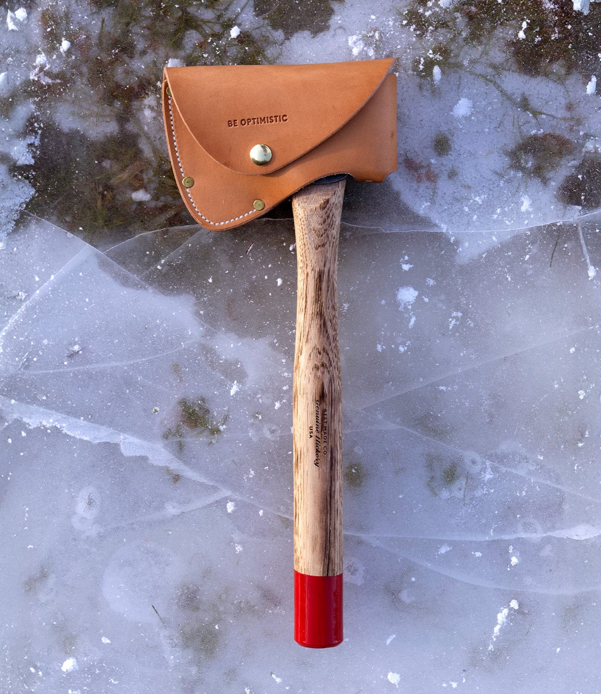 The Spark (Straight-Hold Edition) by Best Made Co. is a high-carbon steel hatchet with an Appalachian hickory handle, red-painted tip, and brass-riveted sheath; its leather blade guard reads BE OPTIMISTIC as it rests on cracked ice.