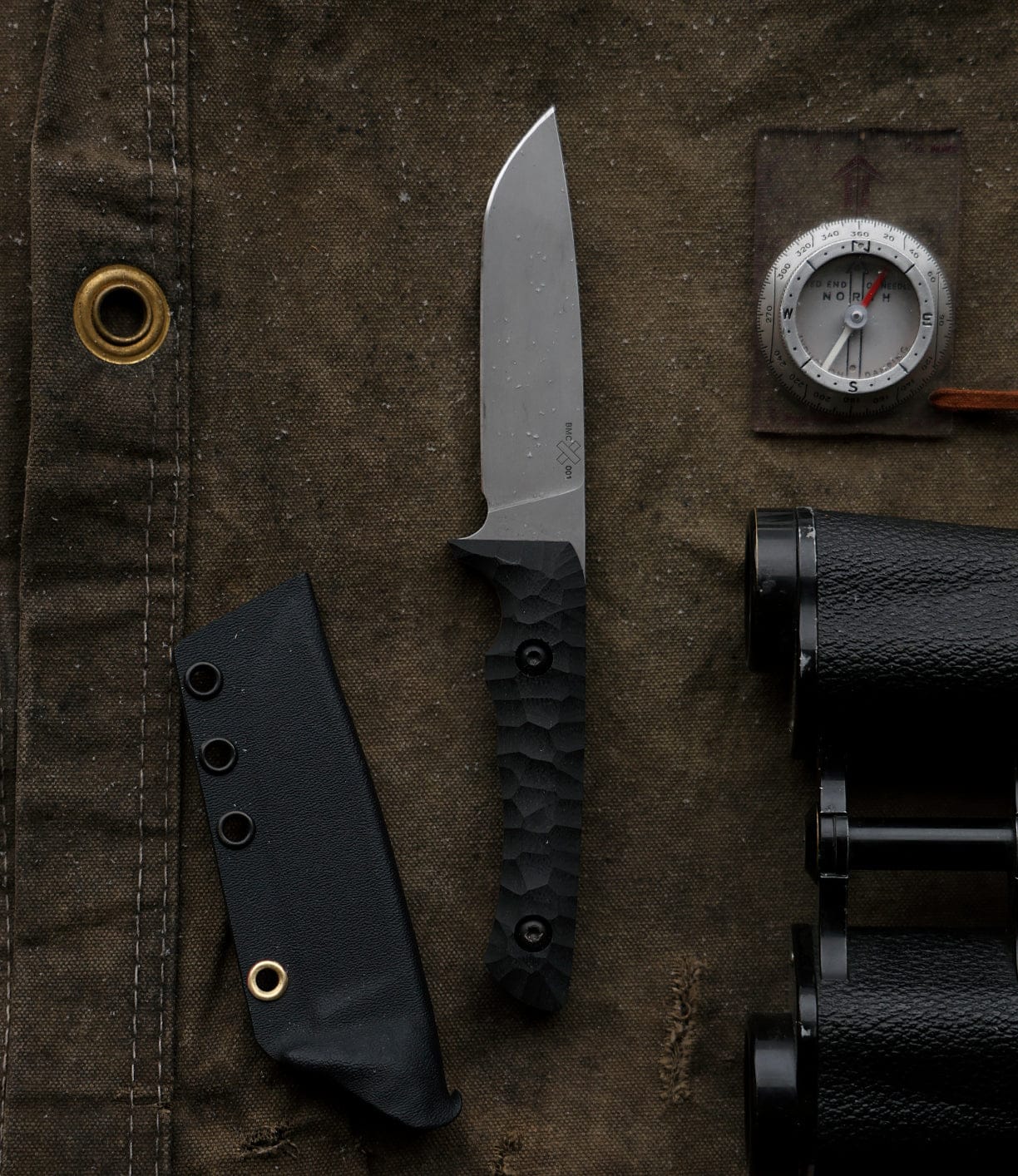 The Best Made Co NWBW Field Knife (BMC Edition) with a black textured handle and sheath lies on brown canvas next to a compass and partial black binoculars.
