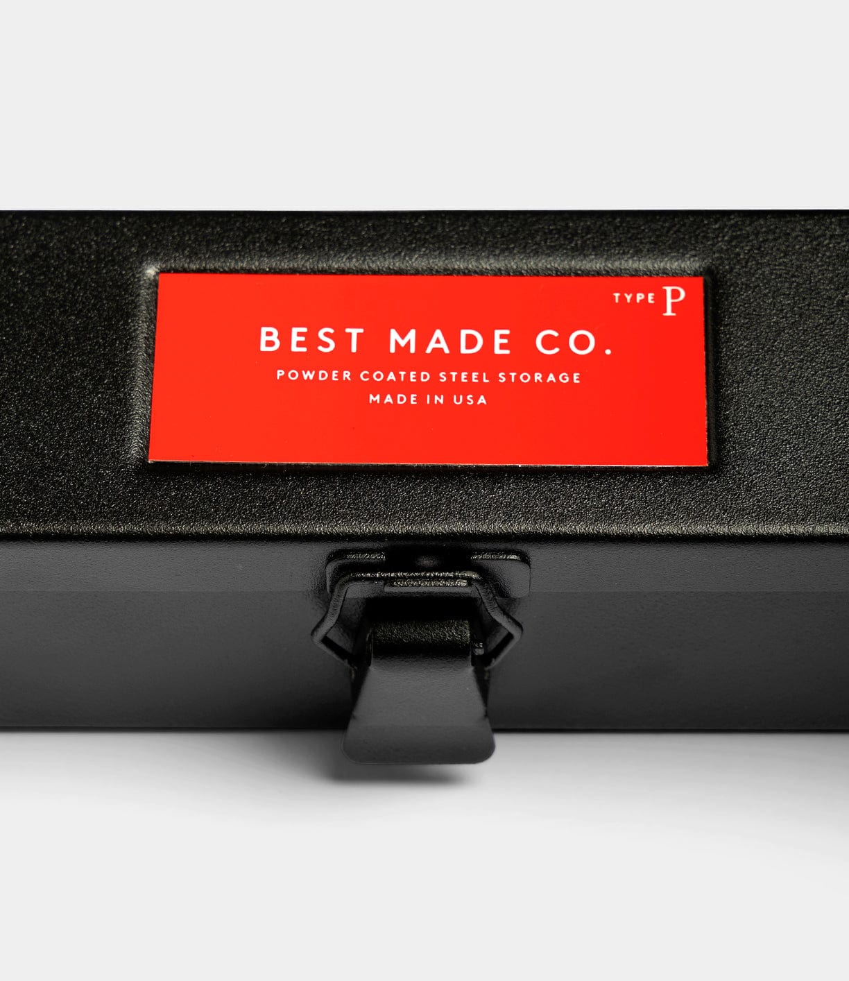 A close-up of a black metal Type P Strongbox, Go Slow Edition by Best Made Co., with a latch and a red label reading Best Made Co. Powder Coated Steel Storage Made in USA.