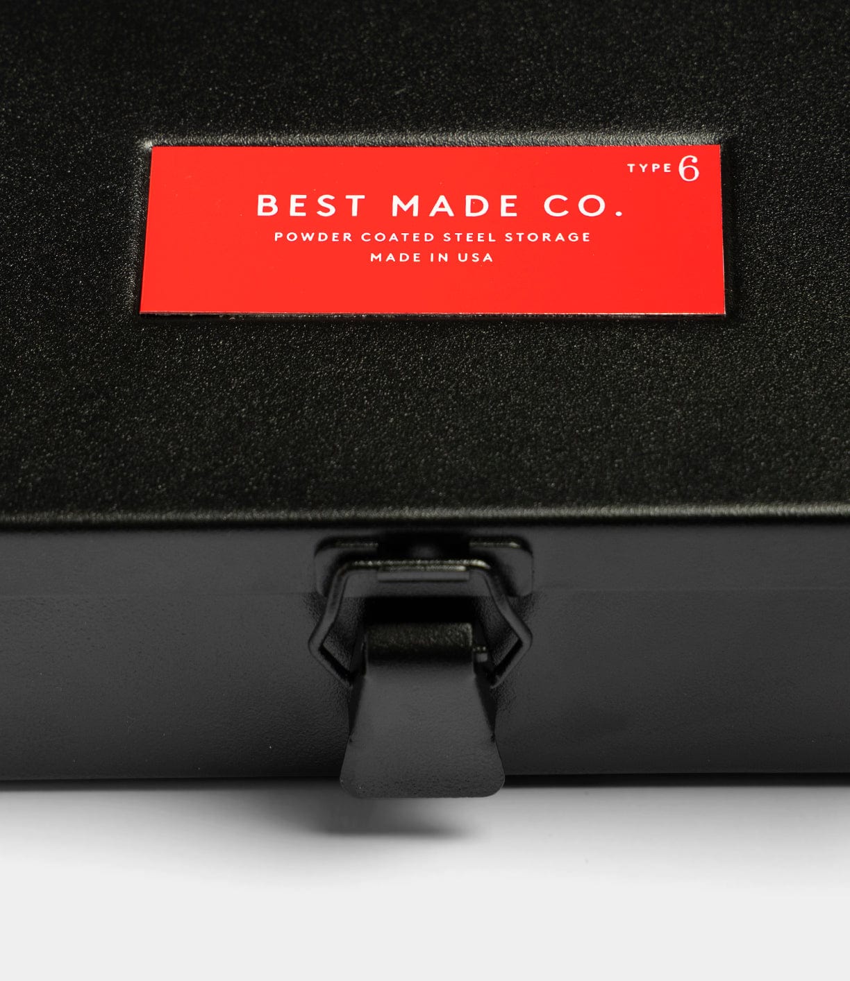 A close-up of a black metal case with a latch and a red label reading Best Made Co. Type 6 Strongbox, Go Slow Edition, Powder Coated Steel Storage, Made in USA.