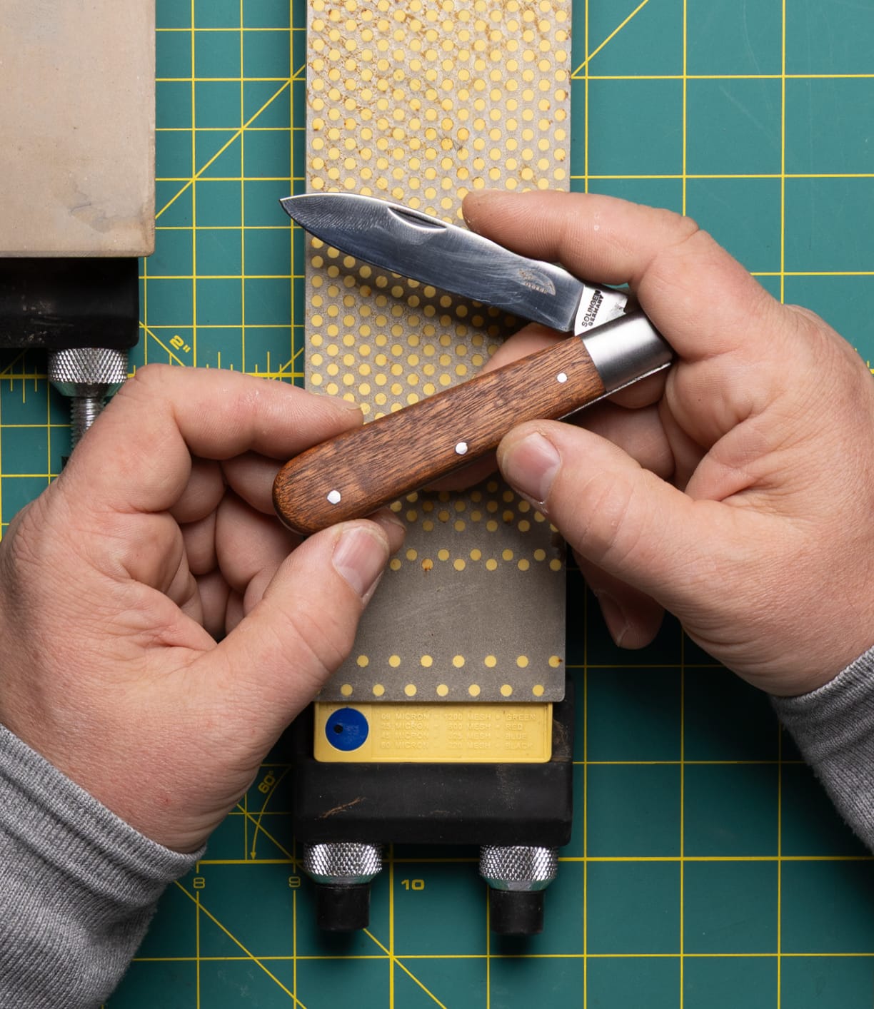 A person sharpens a Best Made Co. Otter-Messer Pocket Knife with a sharpening stone on a green cutting mat, both hands visible holding the knife and stone.