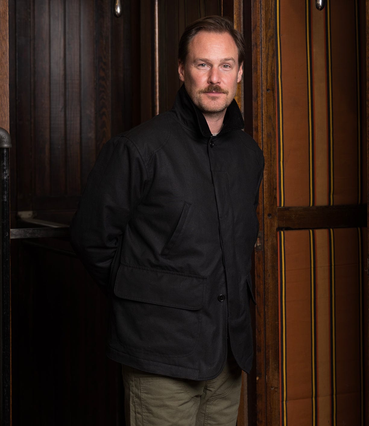 A man with light brown hair, mustache, and beard stands indoors in front of a wooden wall, wearing Best Made Co.'s Wool-Lined Waxed Field Jacket in Black and olive green pants, with his hands in his pockets.