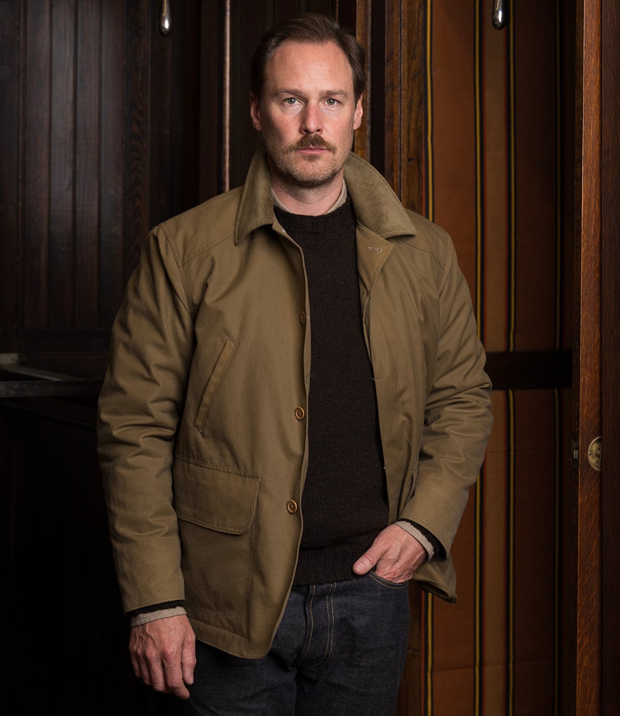 A man with light brown hair and a mustache stands indoors against wood paneling, wearing the Best Made Co. Wool-Lined Waxed Field Jacket in Tan over a dark sweater and jeans, one hand in his pocket.