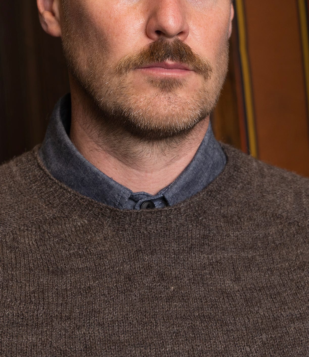 A close-up of a man's lower face and neck with a light brown mustache and beard stubble. He wears a blue collared shirt under the Best Made Co. 5-Gauge Shetland Sweater, offering lightweight warmth.