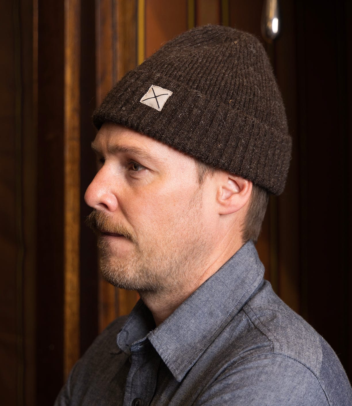 A man with light facial hair wears the Best Made Co Shetland Cap of Courage and a blue button-up shirt, standing in profile against a wooden background.