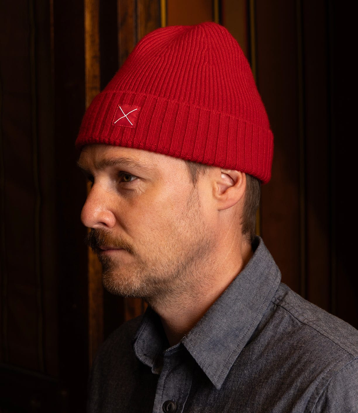 A light-skinned man with a short beard wears a bright red Best Made Co Cap of Courage lambswool cap featuring a white X on the side and a gray button-up shirt, standing indoors against a dark background.