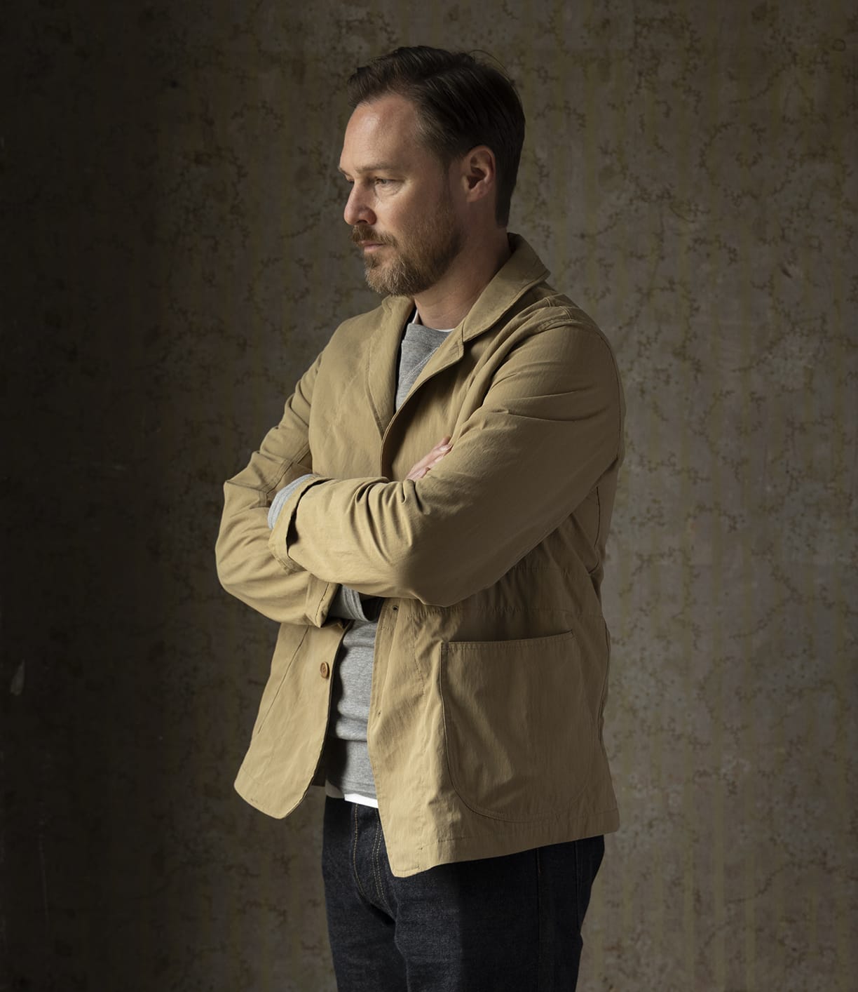 A man with light brown hair and a beard stands with arms crossed, wearing the Best Made Co Possibilities Jacket Tan over a gray shirt and dark denim jeans, looking left against a textured brown background.
