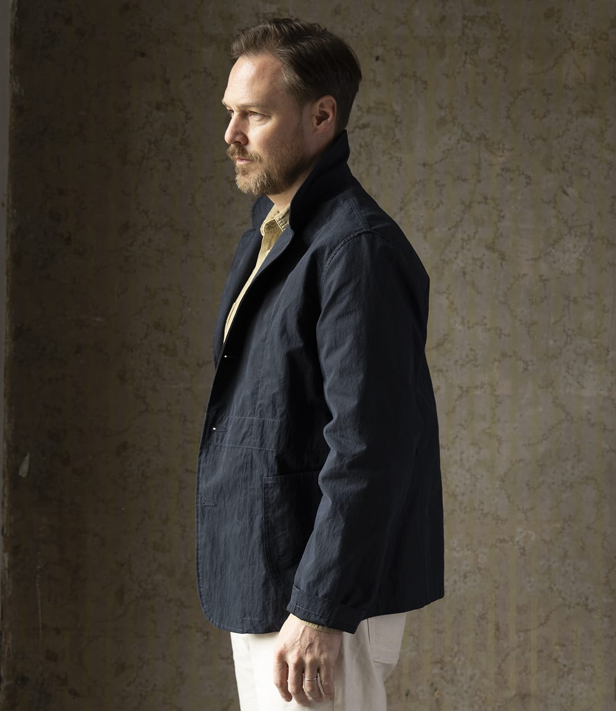 A man with light brown hair and a beard stands in profile against a textured wall, wearing the Best Made Co Possibilities Jacket Navy over a beige shirt and white pants.