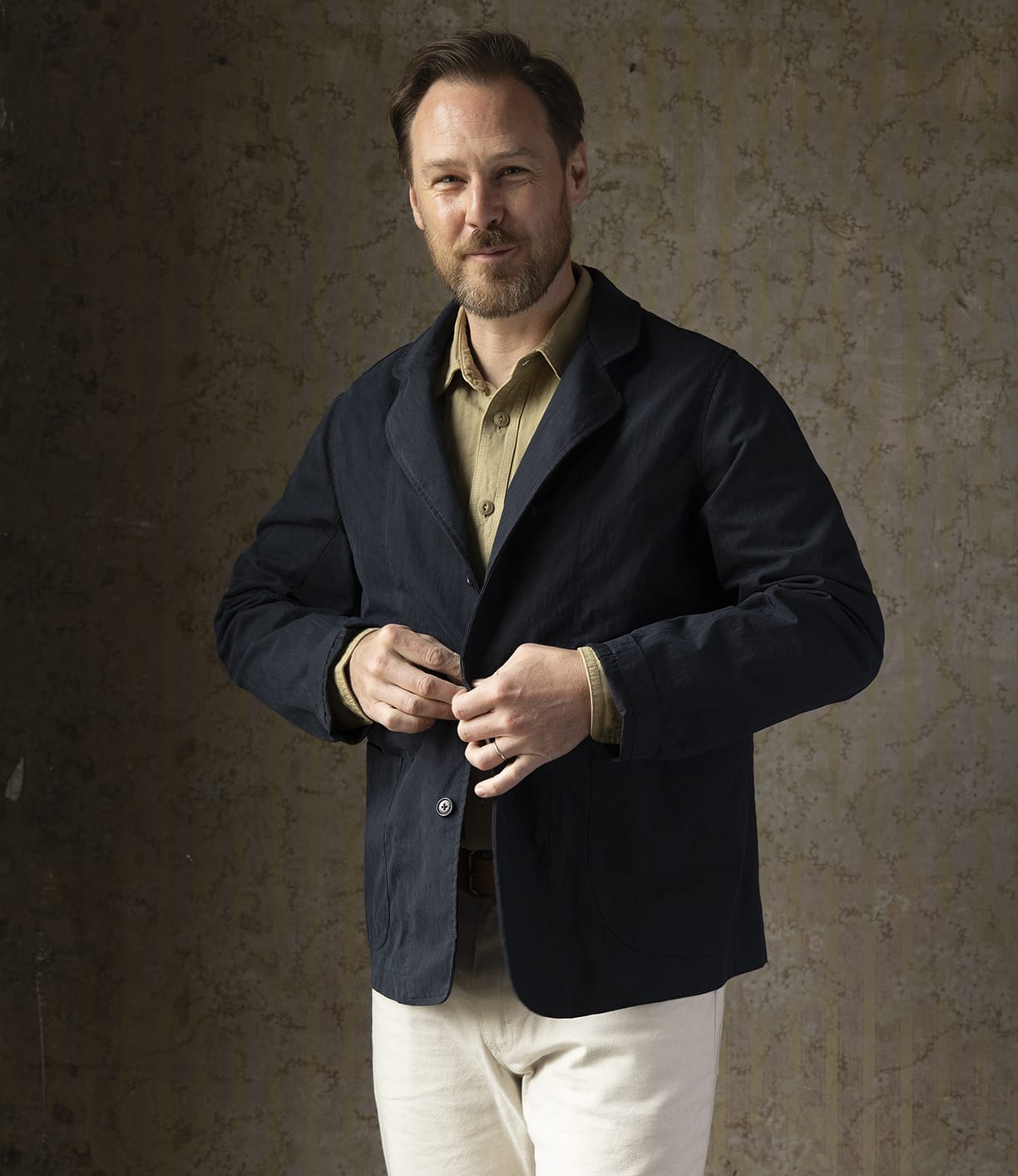 A man with light brown hair and a beard stands against a textured wall, wearing the Best Made Co Possibilities Jacket Navy over a beige shirt and white pants, looking at the camera while buttoning his jacket.